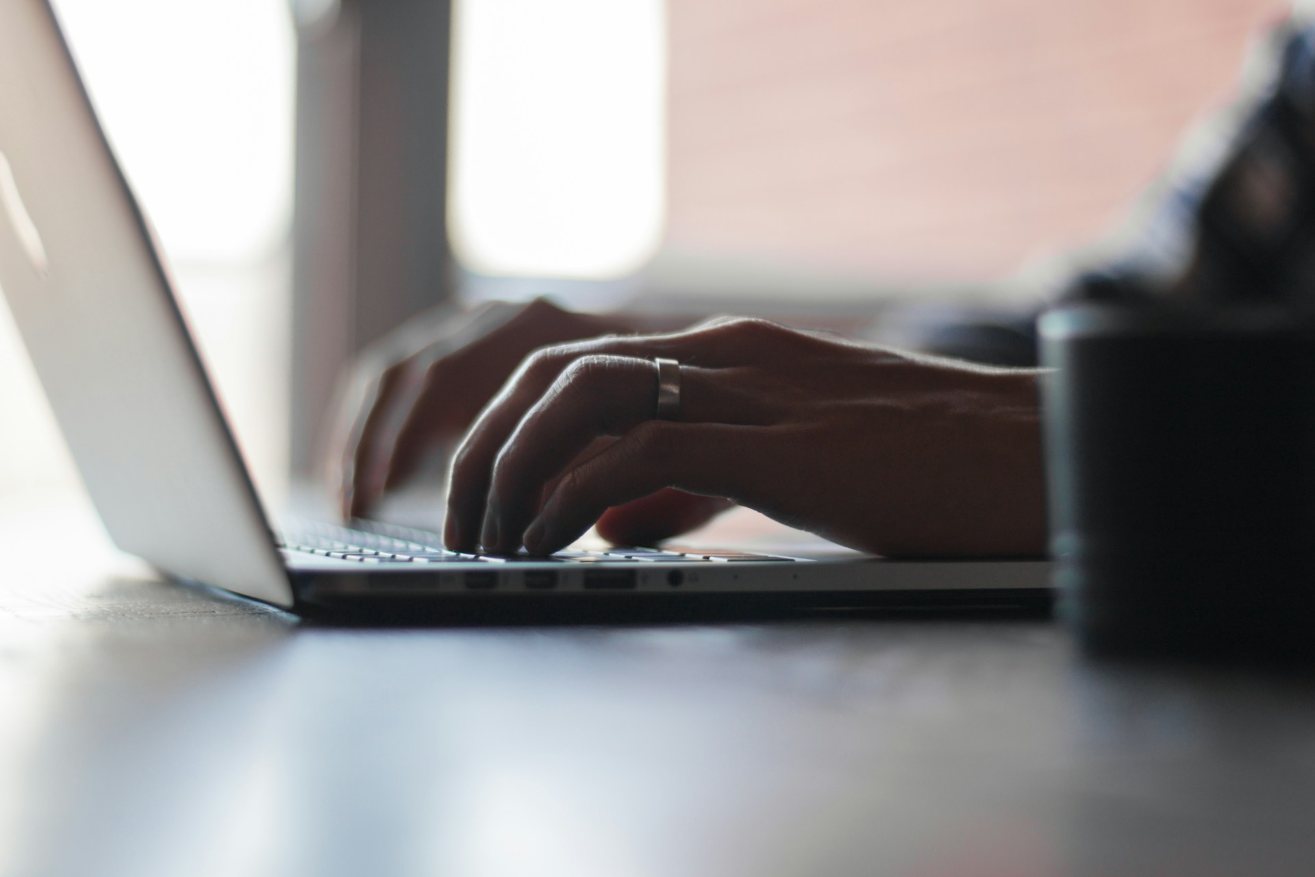 A person typing on a laptop keyboard with both hands in a softly lit workspace with a blurred coffee mug in the foreground.