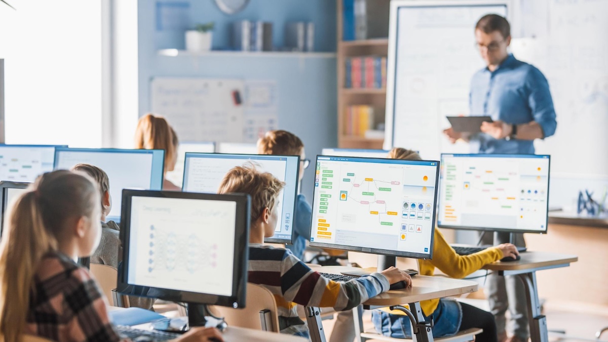 A teacher stands in front of a computer screen with a teacher in front of him